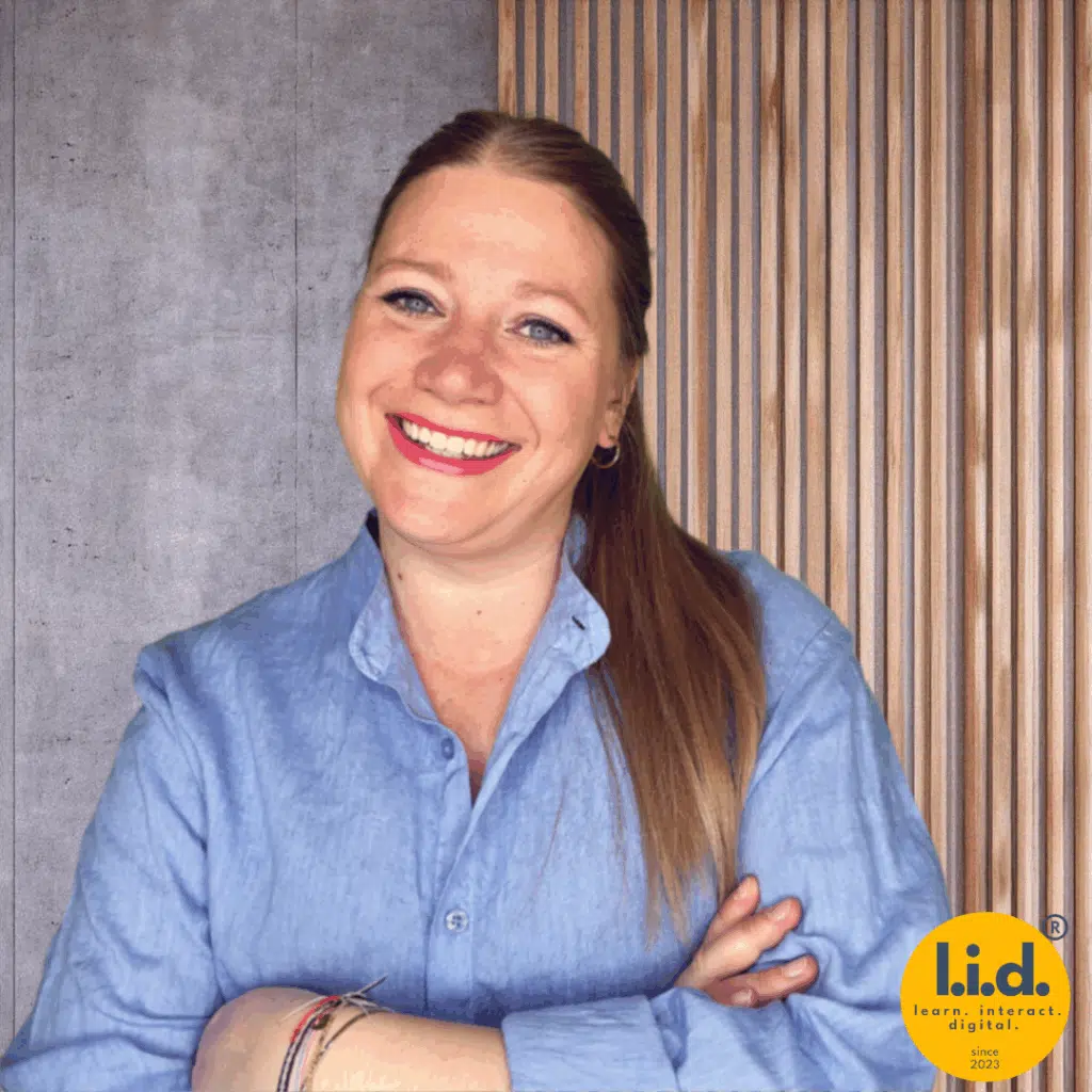 Lea Wurm, lächelnd mit verschränkten Armen, vor einer modernen Wand aus Holzlamellen und Betonoptik. Im Bild unten rechts ist das gelbe Logo von l.i.d. (learn. interact. digital.) sichtbar.